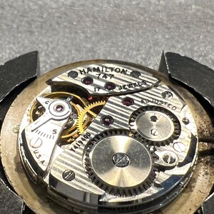 K&ouml;nnte beinhalten: Nahaufnahme eines Vintage-Uhrwerks von Hamilton. Der Uhrenmechanismus zeigt komplizierte Zahnr&auml;der, Juwelen und den Text "HAMILTON 747 JEWELS". Das Uhrwerk ist silberfarben mit goldenen Zahnr&auml;dern und dem Text "U.S.A."