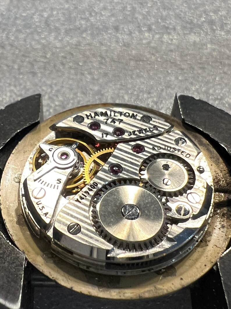 K&ouml;nnte beinhalten: Nahaufnahme eines Vintage-Uhrwerks von Hamilton. Der Uhrenmechanismus zeigt komplizierte Zahnr&auml;der, Juwelen und den Text "HAMILTON 747 JEWELS". Das Uhrwerk ist silberfarben mit goldenen Zahnr&auml;dern und dem Text "U.S.A."