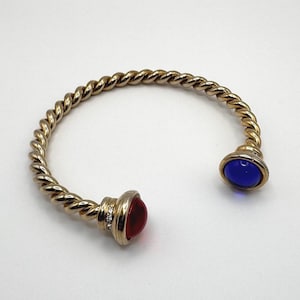 May include: Gold-tone cuff bracelet with a twisted rope design. The bracelet features a red and a blue cabochon stone at each end. The red stone end has three small clear crystals. A stylish piece of jewelry.