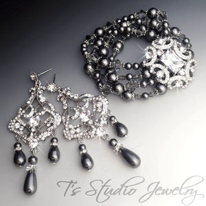 May include: A set of grey pearl and crystal jewellery featuring a bracelet and dangle earrings. The bracelet is made of multiple strands of grey pearls and crystal beads with a large crystal centrepiece. The earrings are made of a crystal design with grey pearl drops.