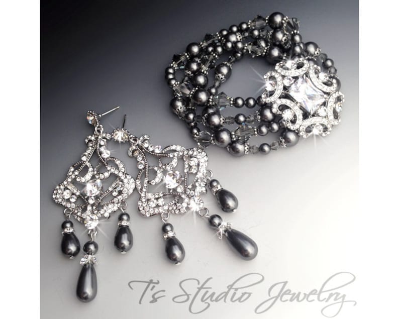 May include: A set of grey pearl and crystal jewellery featuring a bracelet and dangle earrings. The bracelet is made of multiple strands of grey pearls and crystal beads with a large crystal centrepiece. The earrings are made of a crystal design with grey pearl drops.