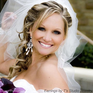 May include: A bride with blonde hair and a white veil smiles for the camera. She is wearing a white strapless dress and dangling pearl and crystal earrings. The text "Earrings by T's Studio Jewelry" is at the bottom of the image.
