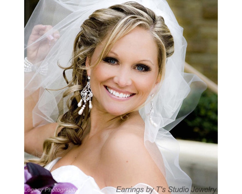 May include: A bride with blonde hair and a white veil smiles for the camera. She is wearing a white strapless dress and dangling pearl and crystal earrings. The text "Earrings by T's Studio Jewelry" is at the bottom of the image.