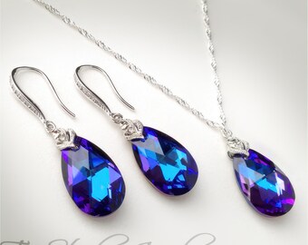 May include: A jewelry set featuring a necklace and earrings. The set includes a silver chain necklace and matching earrings, each adorned with a large, teardrop-shaped, iridescent blue and purple crystal. The earrings have a hook design.