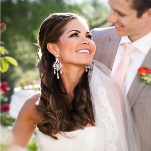 May include: A bride in a white wedding dress with a veil smiles at the groom. She has long brown hair with a side parting and is wearing large crystal chandelier earrings.