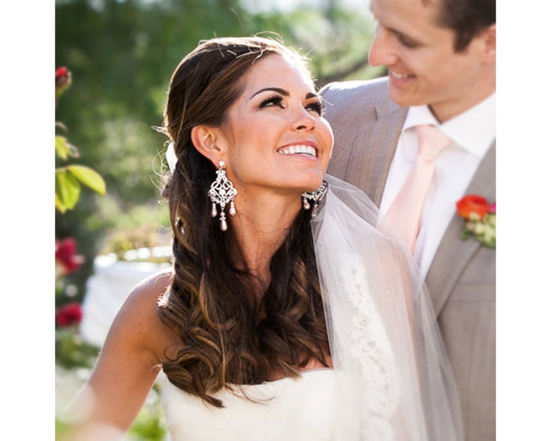 May include: A bride in a white wedding dress with a veil smiles at the groom. She has long brown hair with a side parting and is wearing large crystal chandelier earrings.