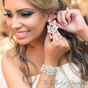 May include: A bride wearing a white dress with a crystal and pearl bracelet and earrings. The earrings are large and dangle with pearl drops. The bride is adjusting her earrings.