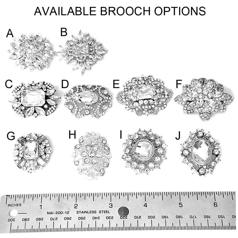 May include: Ten different styles of silver brooches with clear crystal embellishments. The brooches are arranged in two rows of five. A ruler is shown below the brooches for size reference.