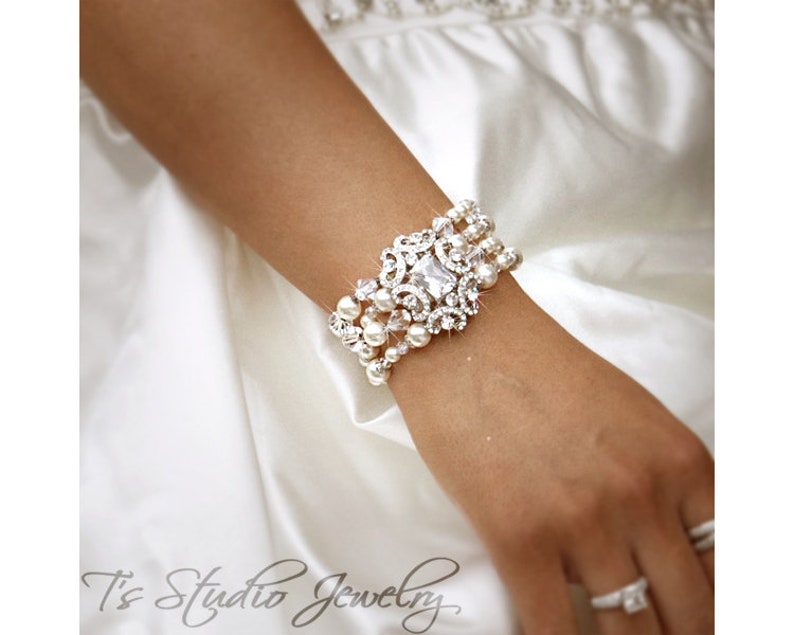 May include: A silver and white crystal and pearl bracelet with a large, ornate, crystal centrepiece. The bracelet is worn on a person's wrist.