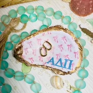 May include: A white oyster shell dish with a pink and white bow pattern and gold trim. The dish has the letters "ADI" written in blue on the front. A pair of gold hoop earrings are sitting in the dish.