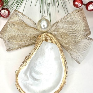 May include: A decorative oyster shell ornament with a gold-painted edge and a pearl-like bead. The ornament is adorned with a gold ribbon bow. The background includes green pine needles and red berries, suggesting a holiday theme.