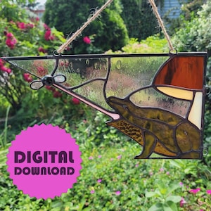 Può includere: Suncatcher in vetro colorato raffigurante una rana stilizzata con la lingua di fuori. L'opera d'arte utilizza tonalità di verde, marrone e ambra, con un bordo nero. Appeso con uno spago. Un adesivo rosa "DOWNLOAD DIGITALE" è visibile.