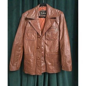 May include: Brown leather jacket with a notched collar, four front pockets, and button closures. The jacket has a tailored fit and a vintage aesthetic. The label reads "Jeffrey Leatherwear".