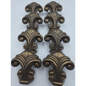 May include: Two matching antique brass-toned metal drawer pulls, each featuring a vertical stack of three ornate fleur-de-lis motifs. The hardware has a dark, weathered finish with curved, symmetrical scrollwork details and a solid, heavy appearance.