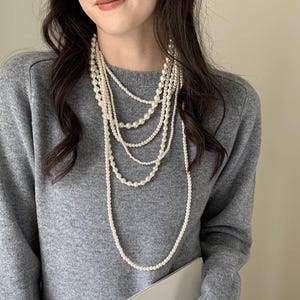 Peut inclure: Un collier de perles à plusieurs rangs de différentes longueurs. Le collier est composé de plusieurs rangs de perles blanches, créant un effet de superposition. Les perles varient en taille, ajoutant de l'intérêt visuel. Le collier est porté sur un pull gris.