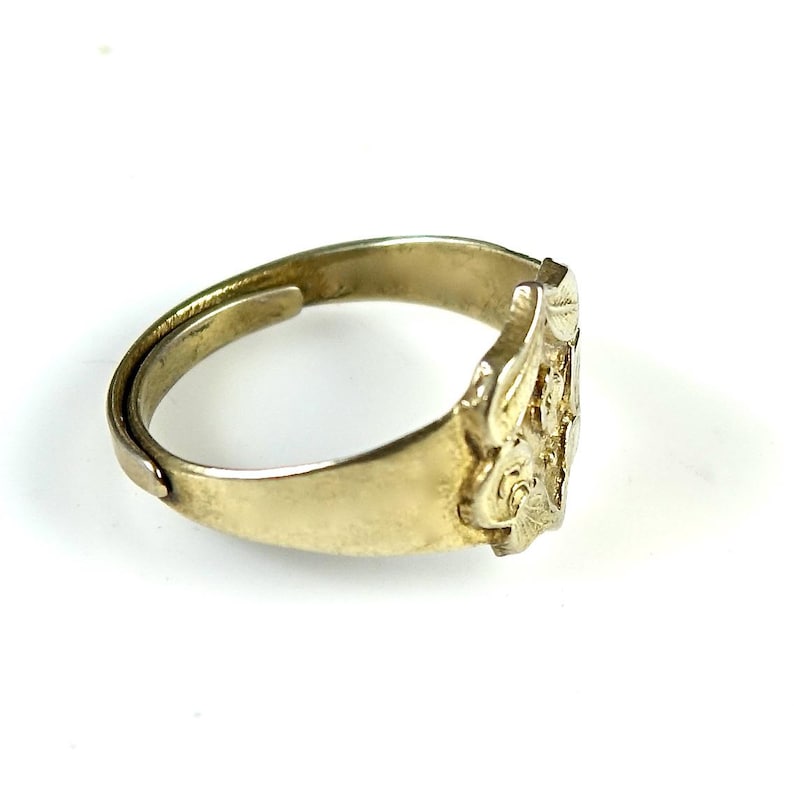 May include: A gold-toned ring with an intricate design on the band. The design features a floral motif with a raised center.