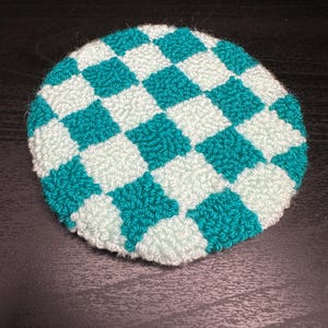 May include: A round, hand-crocheted coaster with a checkerboard pattern. The squares alternate between a teal green and a light mint green. The coaster is made of a soft, textured yarn.