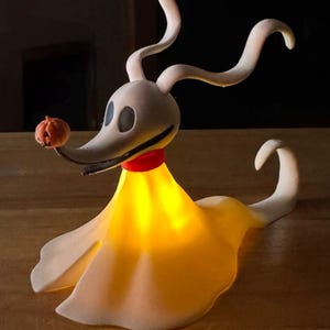 May include: A white, glowing, 3D printed figurine of Zero, the ghost dog from the Nightmare Before Christmas movie. Zero is holding a small orange pumpkin in his mouth and has a red collar around his neck.
