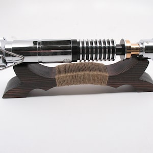 May include: A metallic lightsaber prop with silver and black details, resting on a dark wooden stand. The stand is wrapped with twine. The lightsaber has a cylindrical design with ribbed and smooth sections, and a copper-colored ring.
