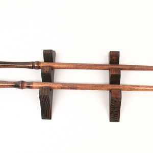 May include: Two wooden wands with a dark brown finish, displayed on a dark brown wooden stand. The wands have rounded handles and tapered shafts. The stand has a simple, geometric design.