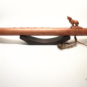 May include: A handcrafted wooden flute with a carved howling wolf figure on top. The flute is a warm brown color, resting on a dark wooden stand. A feather and leather cord are attached. This Native American-style instrument is designed for musical performance.