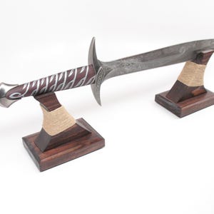 May include: A decorative sword with a silver blade and a handle featuring a burgundy grip with silver accents. The sword rests on two wooden stands with a natural twine accent. The stands are dark brown.