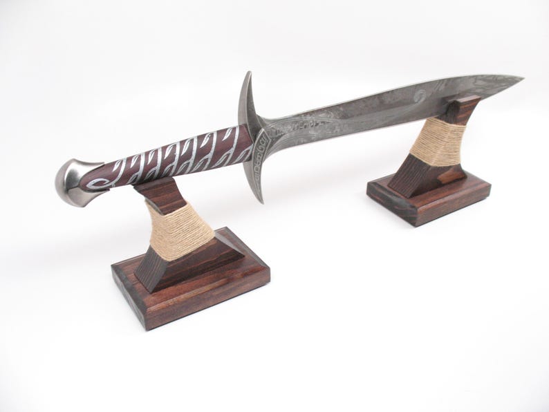 May include: A decorative sword with a silver blade and a handle featuring a burgundy grip with silver accents. The sword rests on two wooden stands with a natural twine accent. The stands are dark brown.