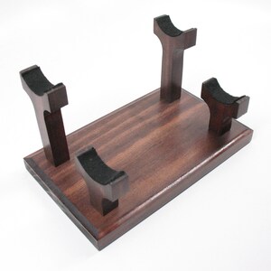 May include: A dark stained wooden display stand with four upright supports, each topped with a black felt cradle. The stand has a rectangular base and is designed to hold an item. The wood grain is visible.