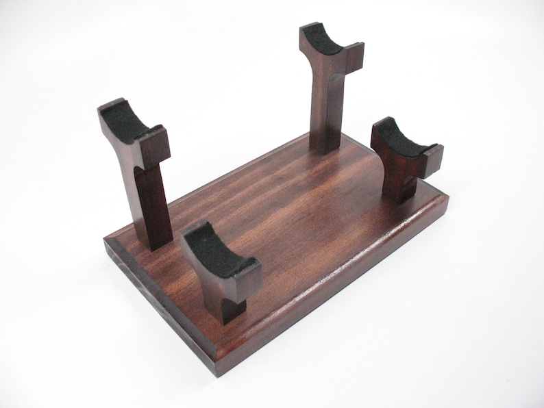 May include: A dark stained wooden display stand with four upright supports, each topped with a black felt cradle. The stand has a rectangular base and is designed to hold an item. The wood grain is visible.