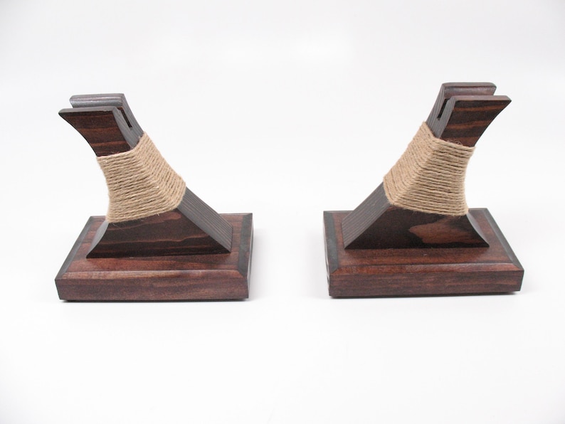 May include: Two dark stained wooden bookends with a square base and curved support. The support is wrapped with natural twine. The bookends are designed to hold books upright on a shelf or desk.