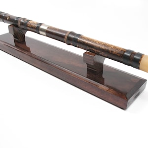 May include: A dark brown bamboo flute with black accents and a light-colored mouthpiece rests on a matching wooden stand. The flute has a silver band and is displayed on a rectangular stand.