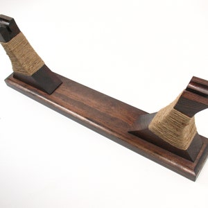 Pode incluir: Um suporte de espada de madeira castanha escura com uma base retangular e dois suportes verticais. Os suportes s&atilde;o envoltos com barbante de cor natural. O suporte foi projetado para segurar uma espada ou item semelhante.