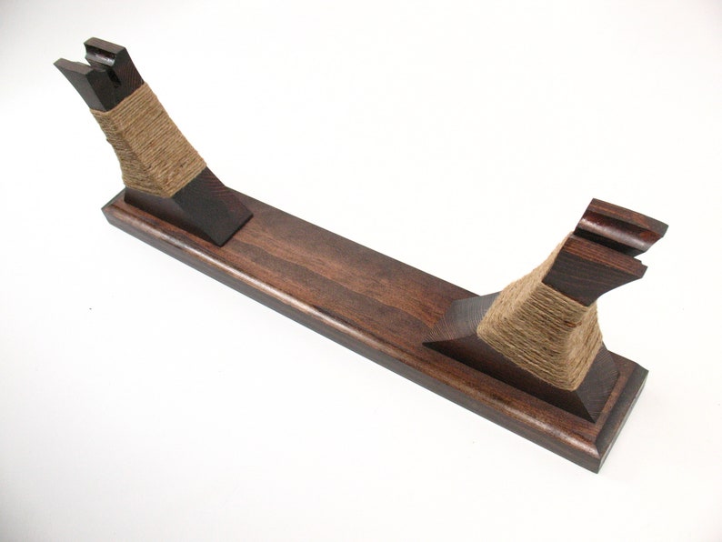 Pode incluir: Um suporte de espada de madeira castanha escura com uma base retangular e dois suportes verticais. Os suportes s&atilde;o envoltos com barbante de cor natural. O suporte foi projetado para segurar uma espada ou item semelhante.