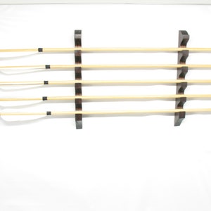 May include: Five wooden arrows with white fletching and black tips are displayed on a dark brown wooden rack. The arrows are arranged horizontally, showcasing their natural wood color and black accents.