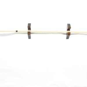 May include: A wooden arrow with a black arrowhead and black accents, displayed horizontally. The arrow is held by two dark brown wooden arrow holders, against a white background. The arrow is made of light-colored wood.