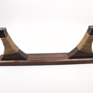Pode incluir: Um suporte de madeira castanha escura com um design curvo. O suporte apresenta uma base retangular e dois suportes verticais envoltos com barbante natural. Os suportes t&ecirc;m uma forma angular &uacute;nica, em fundo branco.