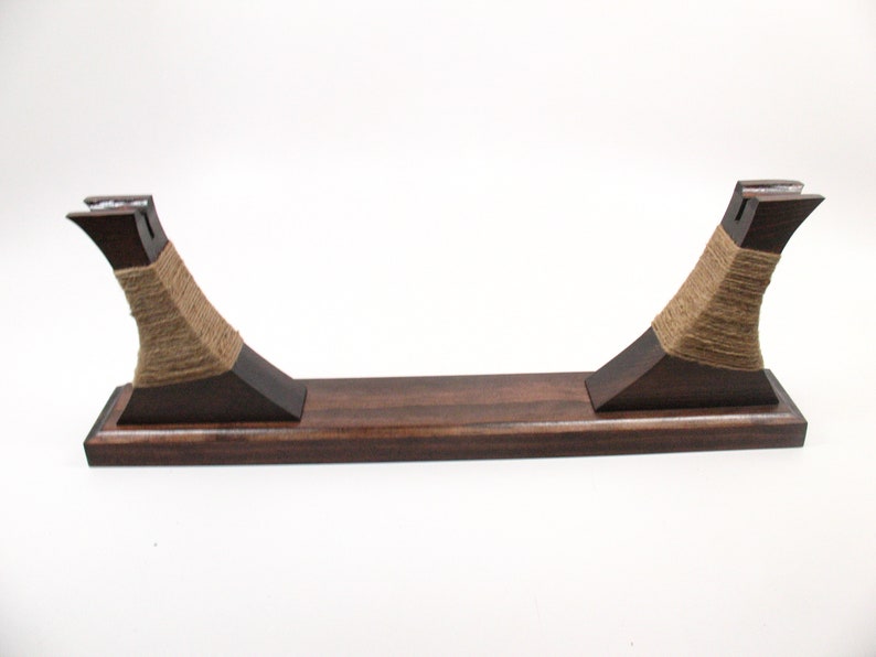 Pode incluir: Um suporte de madeira castanha escura com um design curvo. O suporte apresenta uma base retangular e dois suportes verticais envoltos com barbante natural. Os suportes t&ecirc;m uma forma angular &uacute;nica, em fundo branco.