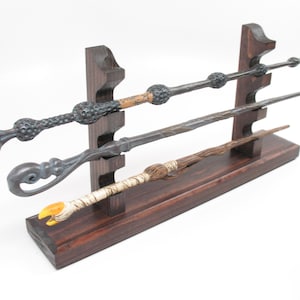 May include: A wooden display stand holding three detailed wands. The wands vary in color and design, with intricate carvings and unique handles. The stand is stained a dark brown, providing a contrasting backdrop.