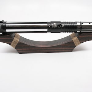 May include: A black and silver lightsaber rests on a dark brown wooden display stand. The lightsaber has a cylindrical design with silver and black accents. The stand is curved and secured with twine.