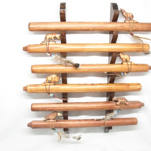 May include: A wooden flute display rack with six flutes. Each flute is a light brown color and has a small animal carving and feathers attached. The rack is made of dark brown wood and is mounted on a white background.