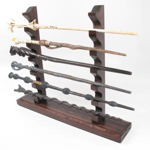 May include: A dark brown wooden wand display stand holding six different wands. The wands vary in color and design, with some featuring intricate carvings and unique shapes. The stand is made of dark wood and has a tiered design.