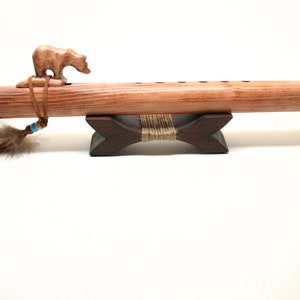 May include: A handcrafted wooden flute with a carved bear figurine on top. The flute is brown with a feather and bead embellishment. It rests on a dark wooden stand, showcasing Native American craftsmanship.