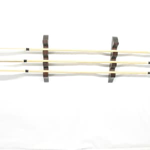 May include: Three arrows with white fletching and black tips are displayed on a dark wooden rack. The arrows have a natural wood shaft with black bands near the fletching and the tip. The rack is mounted on a white background.