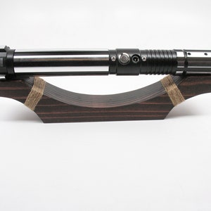 May include: A silver and black lightsaber rests on a dark brown wooden display stand. The lightsaber has a metallic finish with a black handle and a silver blade. The stand is curved and secured with twine.