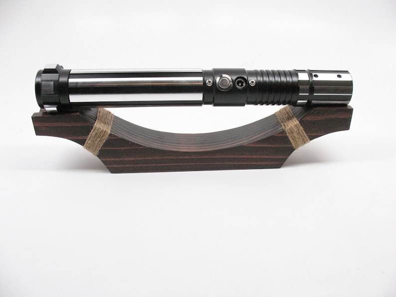 May include: A silver and black lightsaber rests on a dark brown wooden display stand. The lightsaber has a metallic finish with a black handle and a silver blade. The stand is curved and secured with twine.