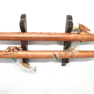 May include: Two wooden Native American flutes, displayed on a dark brown wooden stand. The flutes are a warm brown color, with decorative carvings and feather accents. One flute features a carved eagle.