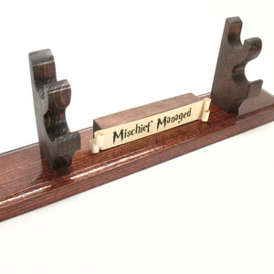 May include: A wooden wand display stand with a dark stain and a glossy finish. The stand features two upright supports with curved cutouts and a central scroll with the words "Mischief Managed".