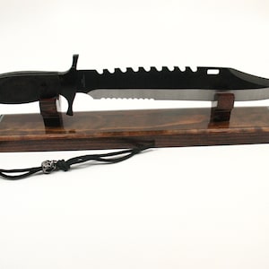 May include: A large black knife with a serrated edge and a pointed tip, displayed on a wooden stand. The knife has a black handle and a lanyard with a skull bead. The stand is a dark brown color.