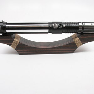 May include: A black and silver lightsaber rests on a dark brown wooden display stand. The lightsaber has a cylindrical design with silver and black accents. The stand is curved and secured with twine.