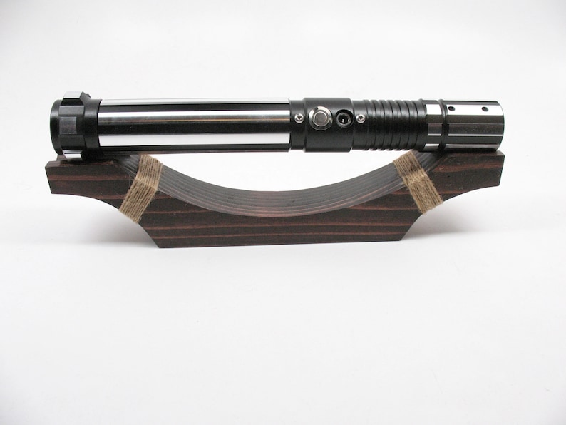 May include: A black and silver lightsaber rests on a dark brown wooden display stand. The lightsaber has a cylindrical design with silver and black accents. The stand is curved and secured with twine.